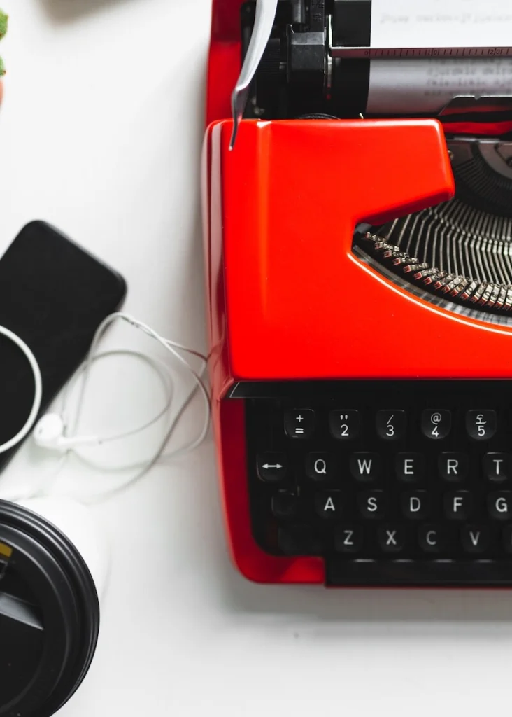 Image of the left side of a red manual typewriter, looking down. Some assorted modern electronics and wires are scattered to the side with a white background.